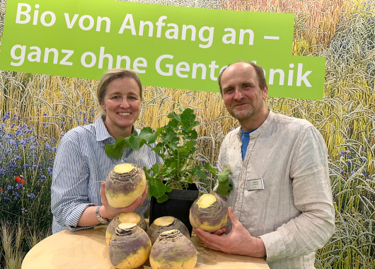 Susanne Schoof und Sebastian Vornhecke halten aufgeschnittene Steckrüben in der Hand. Sie stehen vor einem Schild mit dem Text darauf: Bio von Anfang an - ganz ohne Gentechnik.